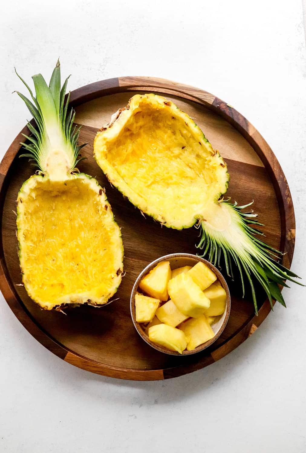 Two pineapple halves hollowed out next to each other on a brown round serving platter with a glass bowl of pineapple chunks in front of the pineapple. 
