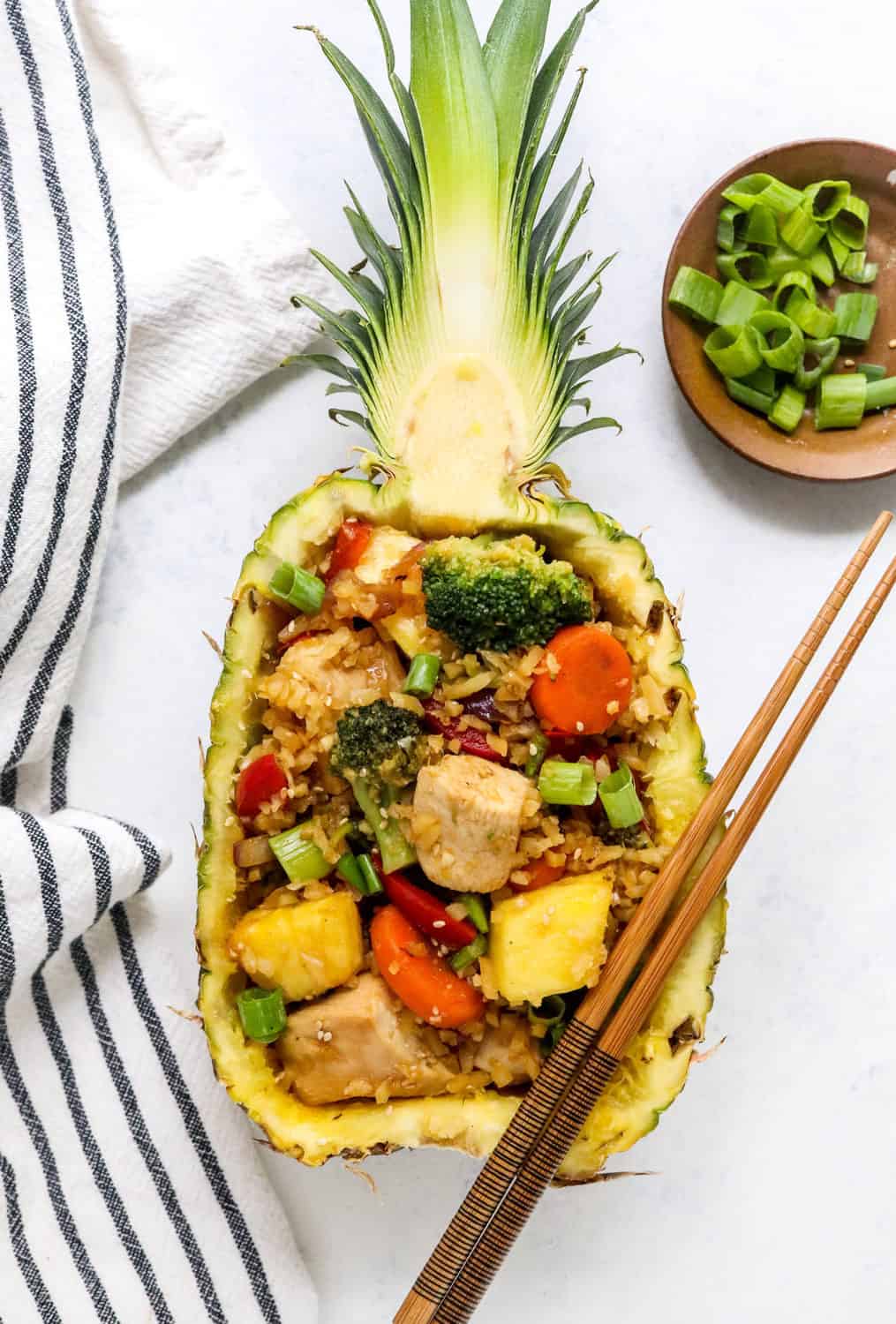 whole half of a pineapple filled with chicken and veggies with a pair of brown chopsticks sitting on the top side of it with a brown bowl of sliced scallions next to the pineapple.
