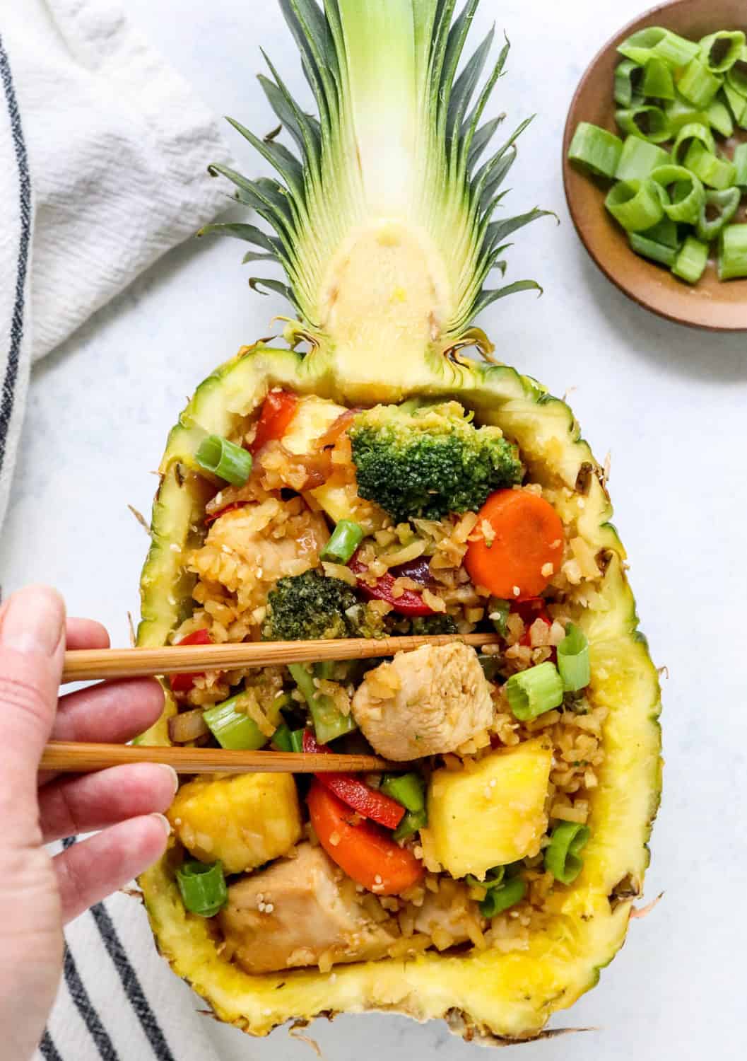 Hand using chopsticks to grab a chunk of chicken out of a pineapple filled with chicken and veggies stir fry. 