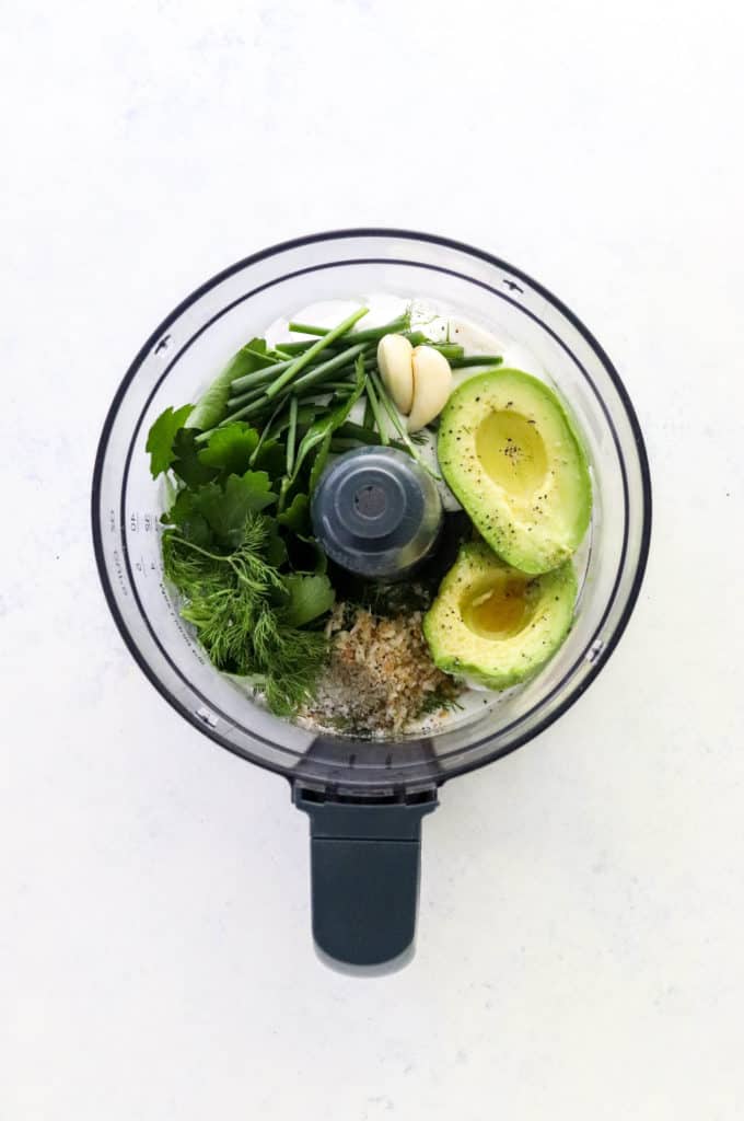 Bowl of a food processor filled with a sliced avocado, fresh herbs, garlic cloves, salt and pepper in it. 