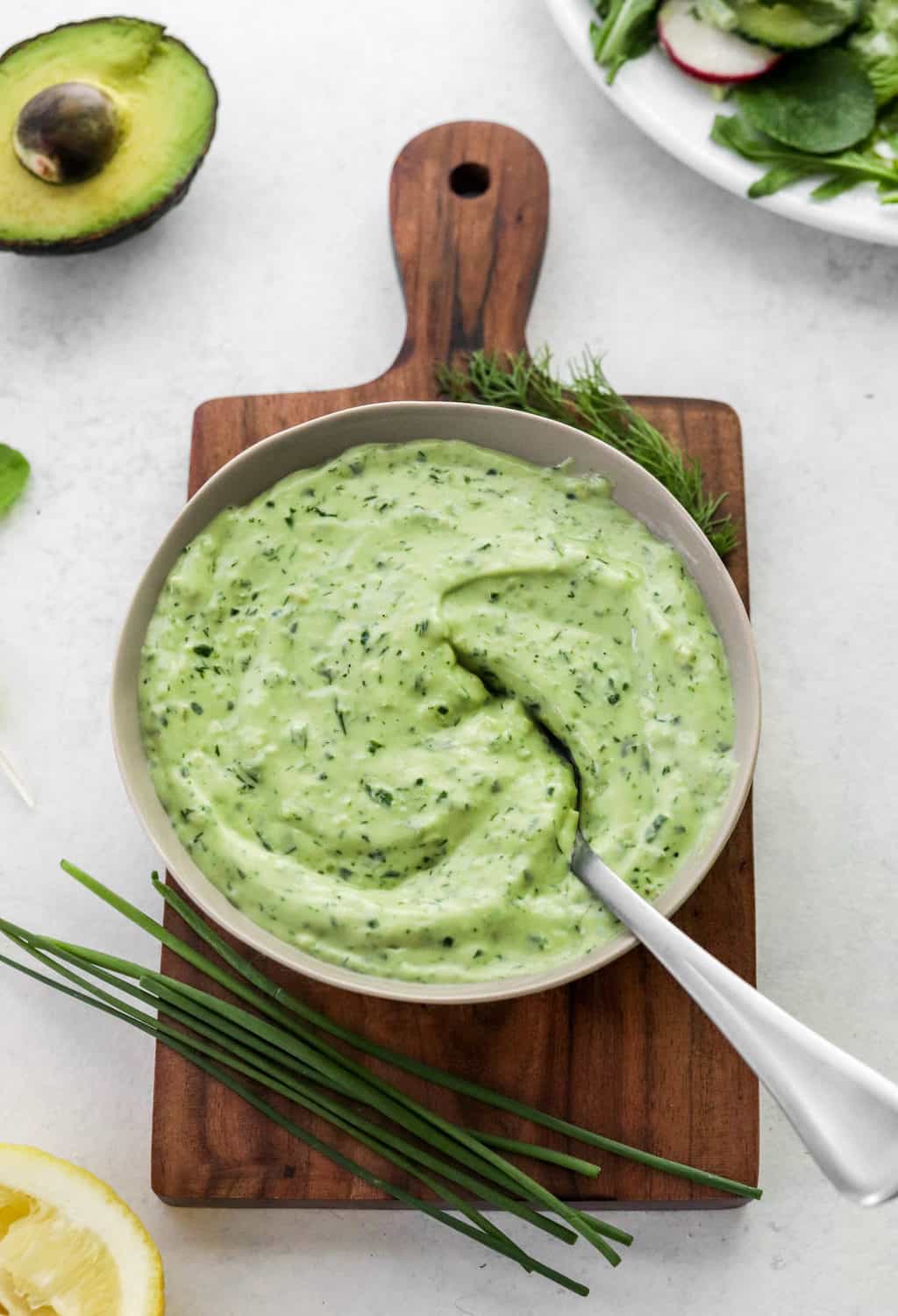 Bowl of green herby dressing with a spoon sticking out of the bowl on top of a small wooden cutting board with chive and dill on the board and a squeezed lemon in front of it. 