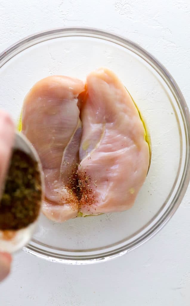Hand pouring spices onto two raw pieces of chicken in a glass bowl
