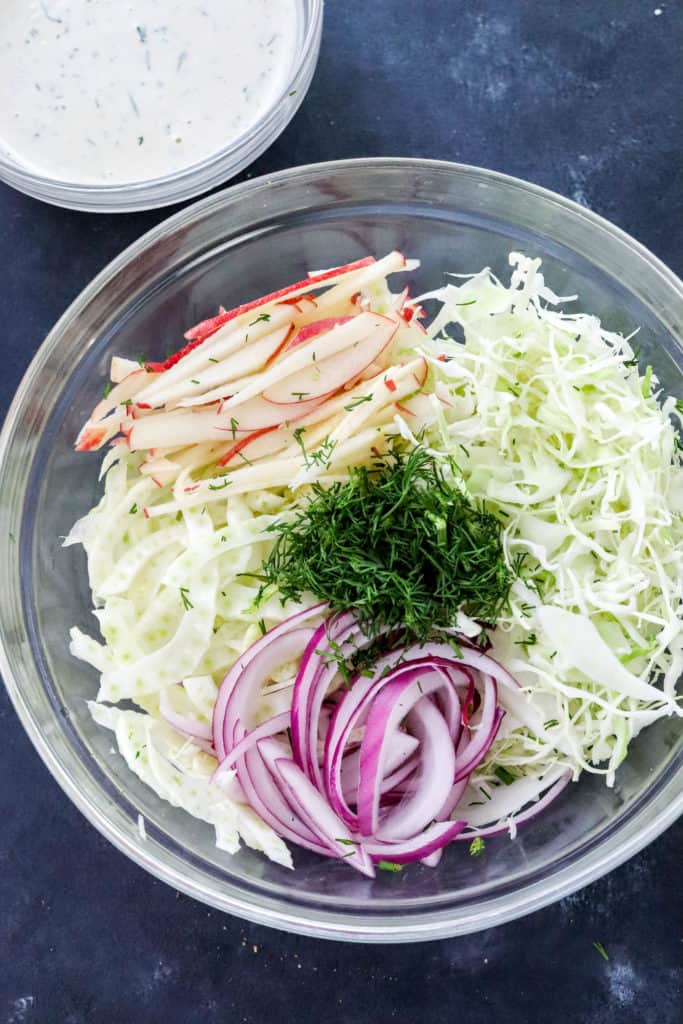 Sliced fennel, shredded cabbage, sliced red onion and dill in a round glass bowl with a bowl of creamy dressing in back of it