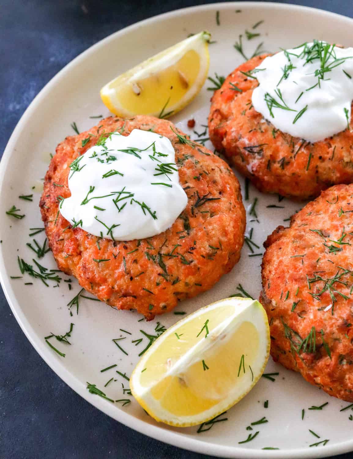 3 round salmon patties on a round plate with some yogurt on top of two of the patties with dill sprinkled on them and lemon wedges next to them. 