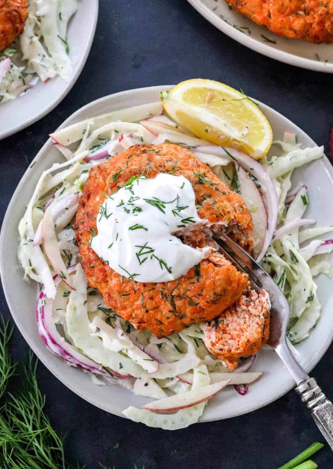 Cooke salmon patty on top of Cole slaw with some greek yogurt and herbs on top of if with a fork cutting through in on a round plate. 