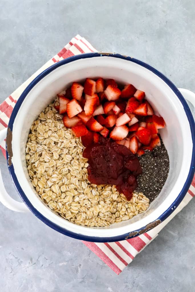 White blue rimmed pot filled with chopped strawberries, jam, oats and chia seeds on top of a red stripped cloth