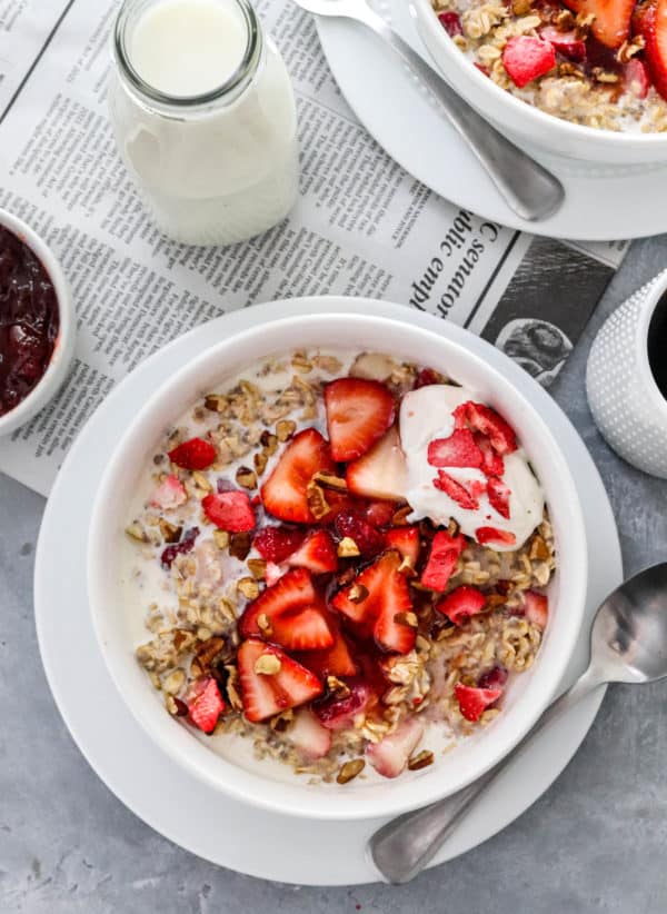 White round bowl on top of a plate filled with creamy oatmeal with glazed chopped strawberries on top of it with a bowl of jam and a cup of coffee next to it