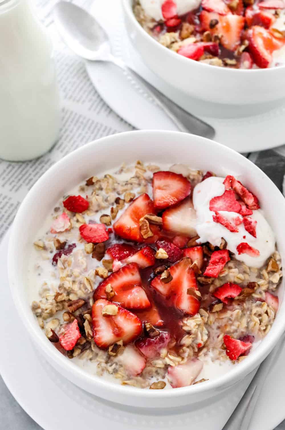 Round white bowl on top of plate filled with creamy oats, sliced strawberries, chopped nuts and yogurt with another bowl behind it. 