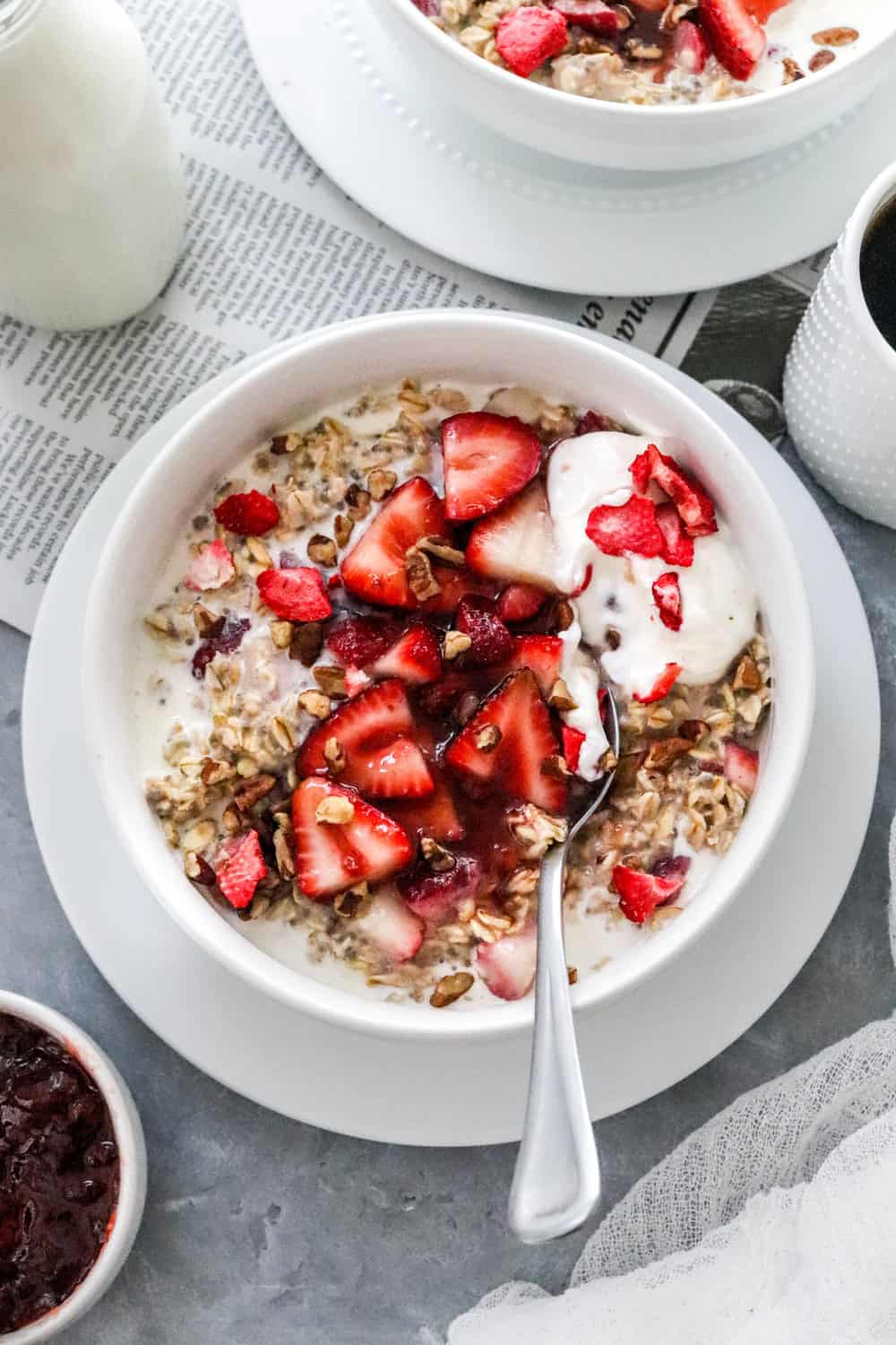 Round white bowl filled with creamy oats topped with sliced strawberries, yogurt and chopped nuts with a spoon in the bowl and another bowl of oats behind it