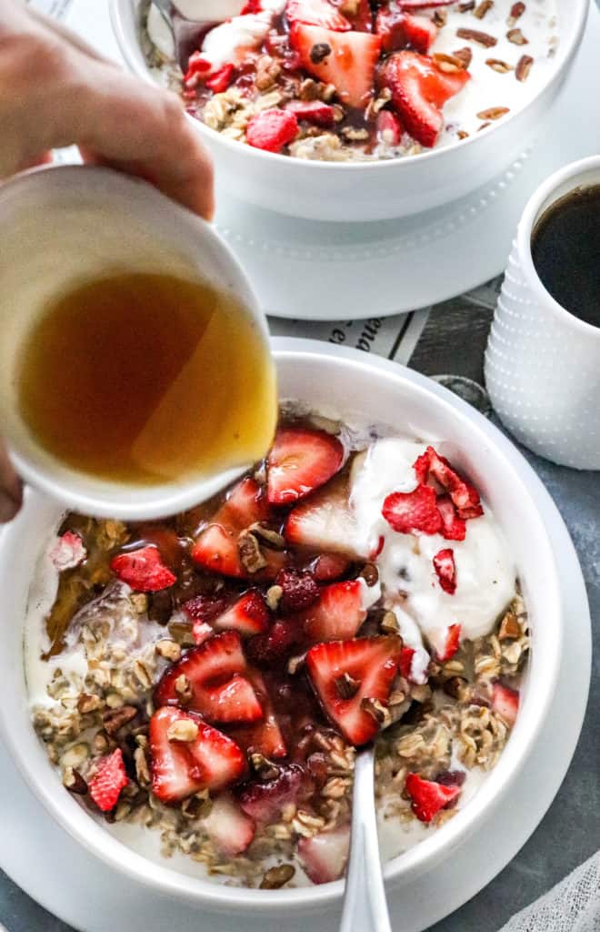 Maple syrup being poured into a bowl if oats and strawberries with a white cup of coffee next to it