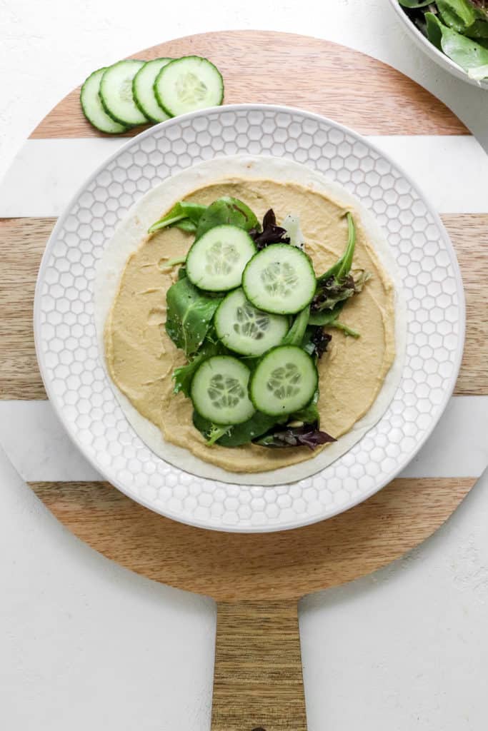 Round wooden cutting board with sliced cucumbers on top of it with a round textured white plate on it that has a tortilla spread with hummus and more greens and sliced cucumbers on top of it