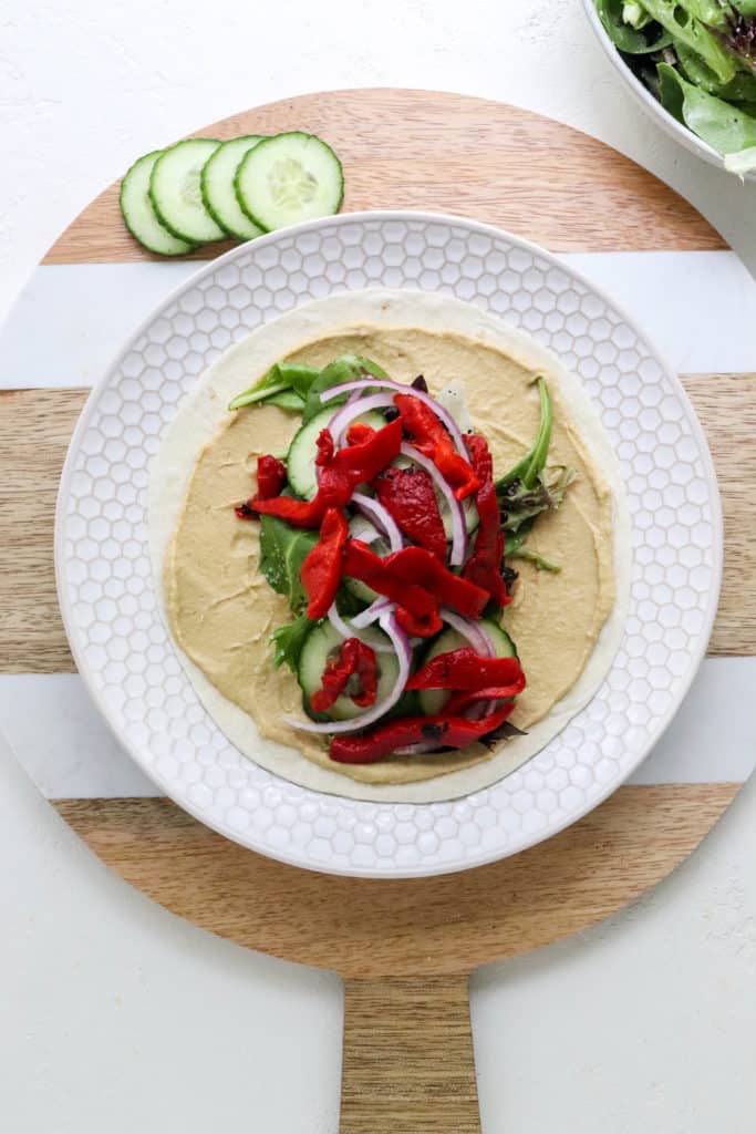 Round textured white plate on top of a round wood cutting board with a white tortillas on the plate topped with hummus and green and red veggies