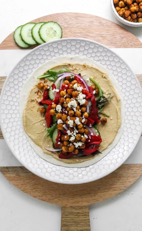 A round cream plate with a round white tortilla on top of it with hummus spread all over it and topped with greens, veggies and crispy chickpeas