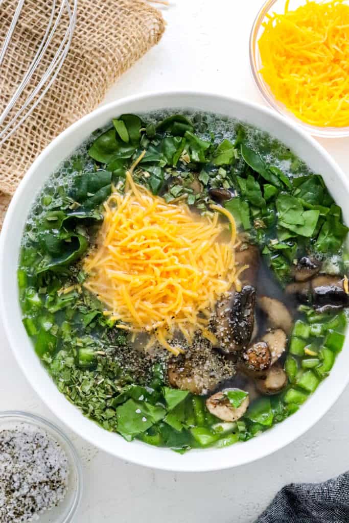 Round white bowl filled with egg whites, spinach, mushrooms and cheddar cheese with a whisk next to it. 