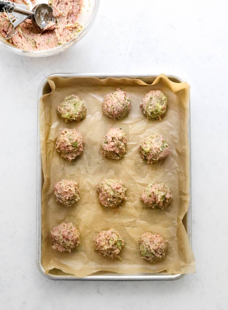 Baking sheet covered in brown parchment paper topped with 12 rolled turkey meatballs with a bowl of the meatballs mixture behind it