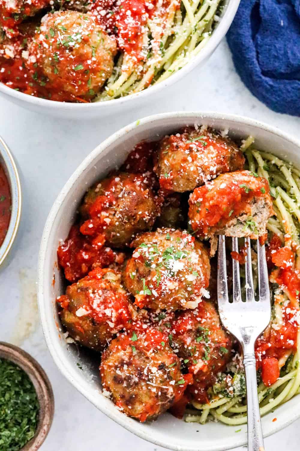 Turkey zucchini meatballs  rolled in tomato sauce on top of spaghetti on a bowl with a fork in the meatballs and a bowl of parsley next to it.