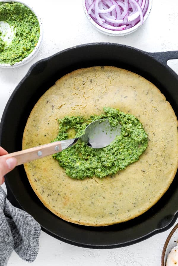 Spoon spreading green pesto all of a pizza crust that is in a round black pan