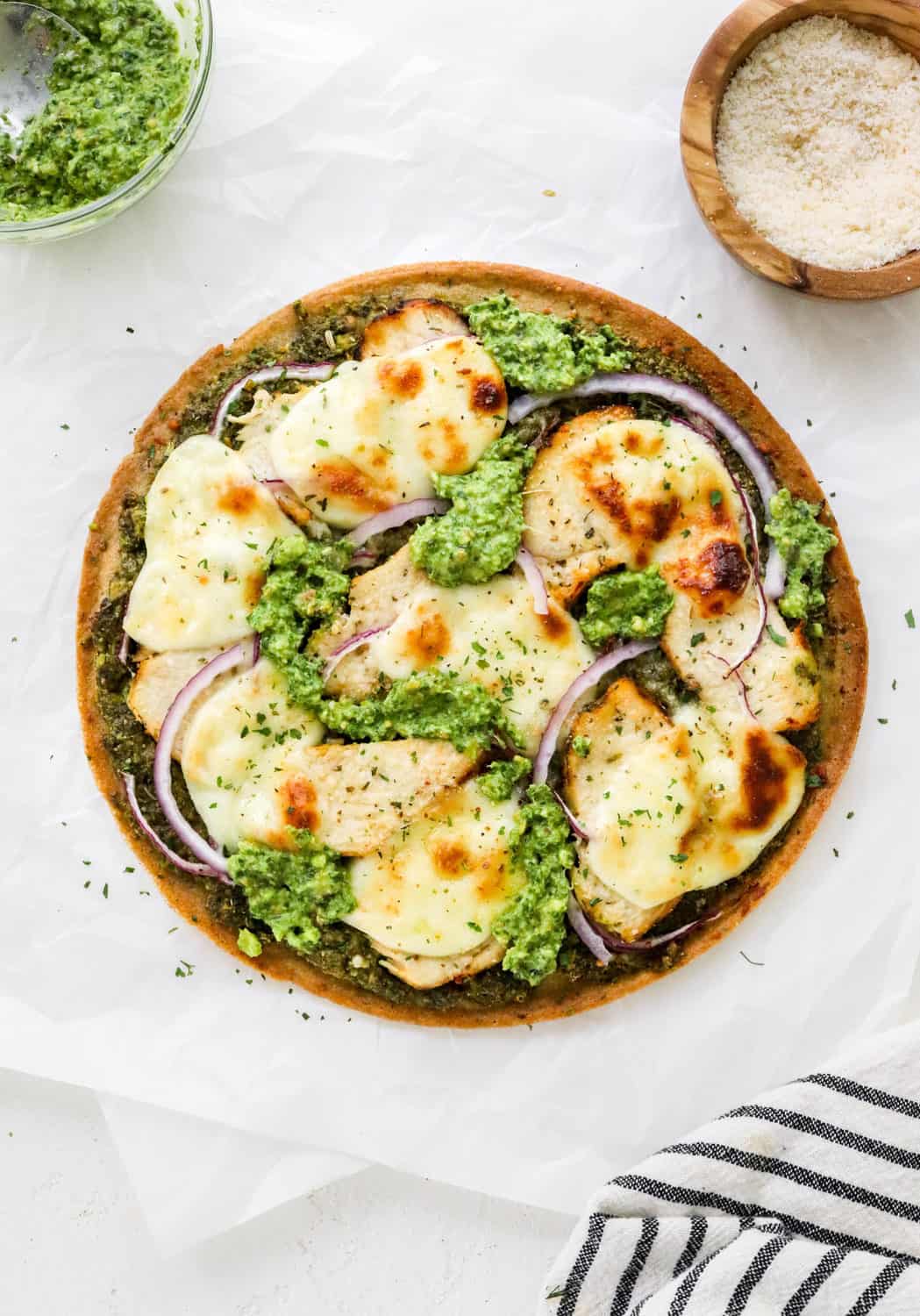 Round thin crust pizza topping with sliced chicken, green pesto and melty cheese on a white surface with more pesto and cheese in bowls behind it. 