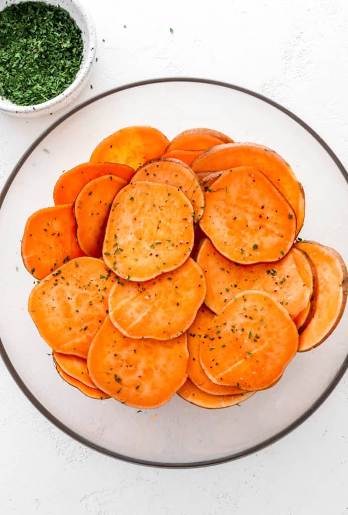 Round glass mixing bowl filled with seasoned sweet potato sliced with a bowl of dried parsley behind it