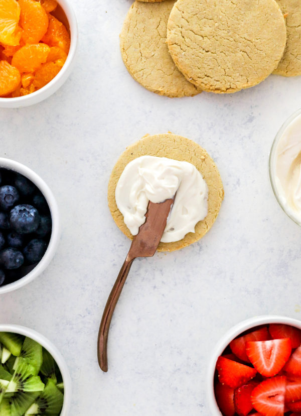 Round sugar cookie with a knife spreading frosting on it with more cookies behind it with bowls of fruit around it