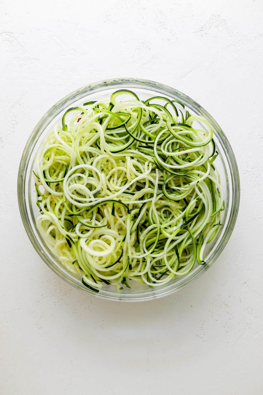 Round glass mixing bowl filled with spiralled zucchini on a white surface. 