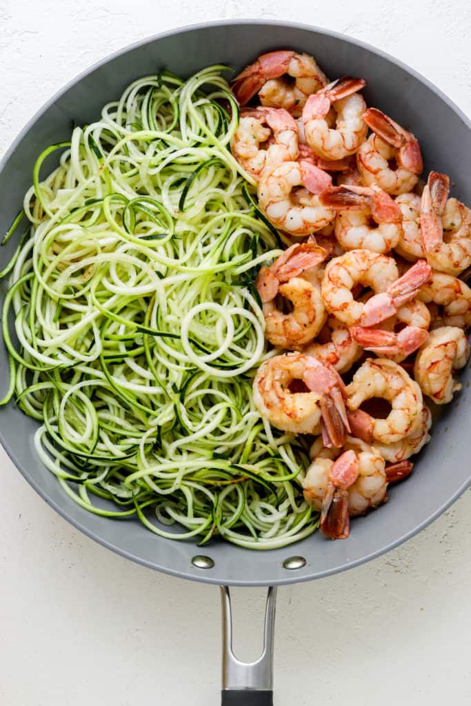 round grey pan with zucchini noodles on one half of the pan and cooked shrimp on the other half of the pan on a white surface