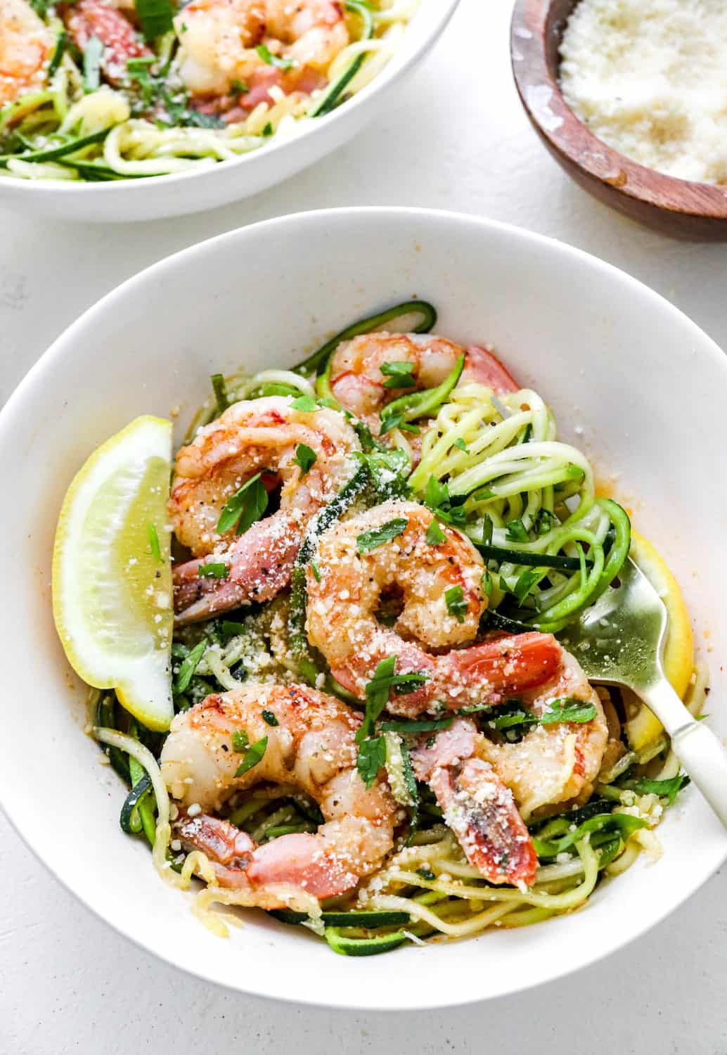 Round white bowl filled with zucchini noodles with cooked healthy shrimp on top with a fork in the bowl with another bowl behind it and a wooden bowl of parmesan cheese behind it. 
