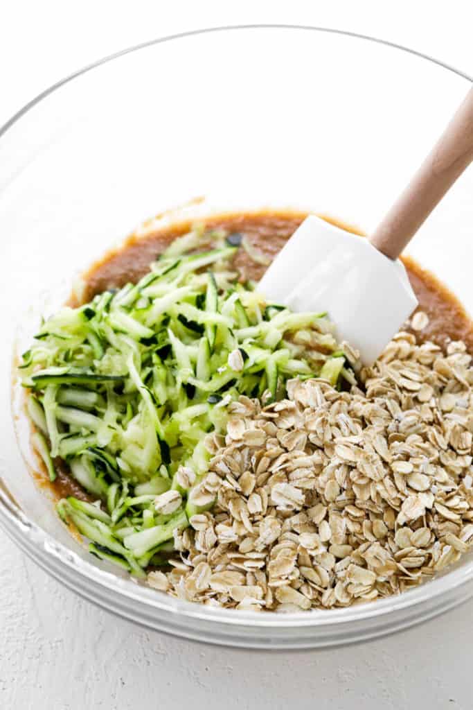 Round glass bowl with brown batter in it with oats and shredded zucchini on top of it with a white spatula in the bowl. 