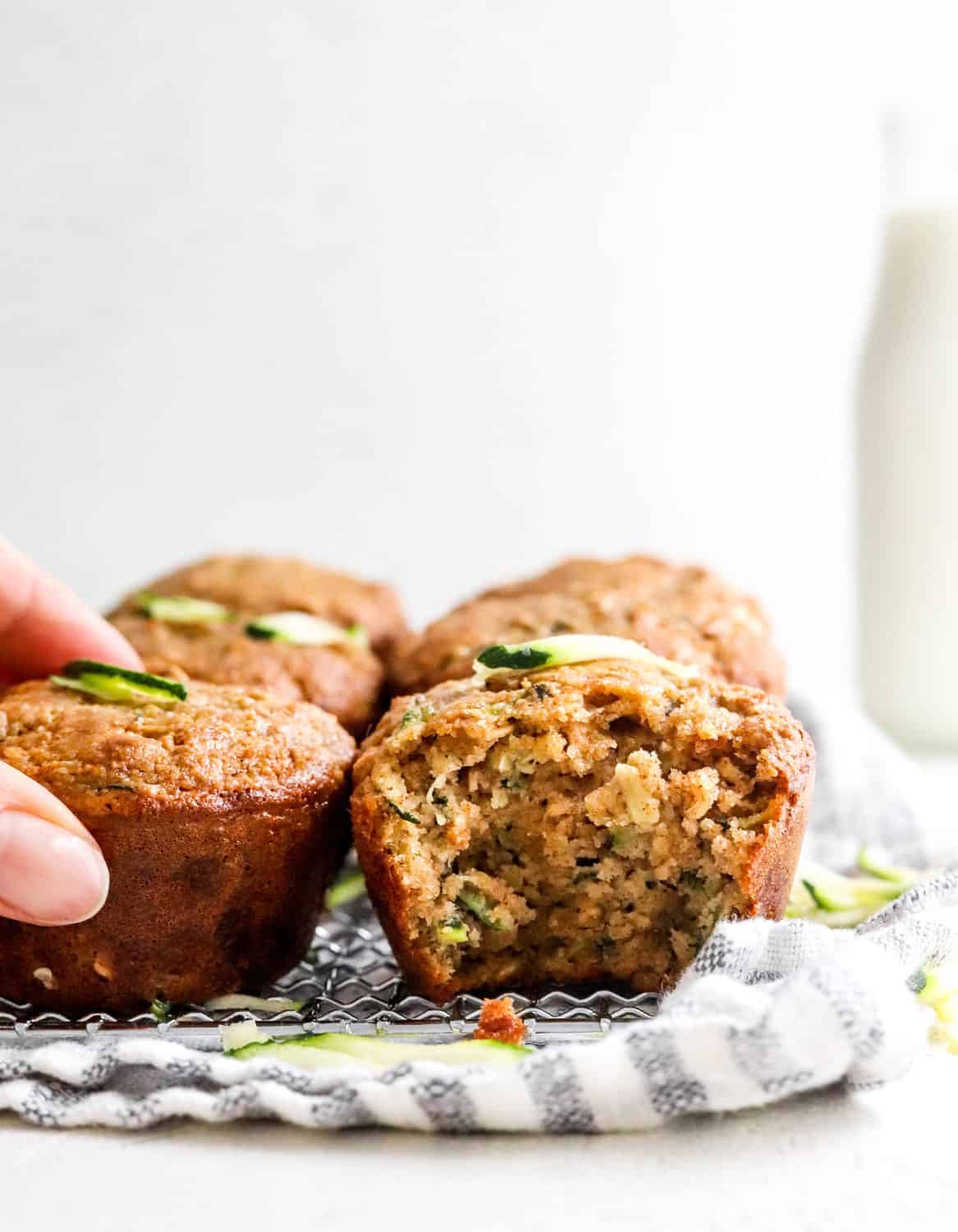 Hand grating a zucchini muffin with more muffins next to it onto of a wore rack with a striped towel under it and pieces of shredded zucchini around it. 