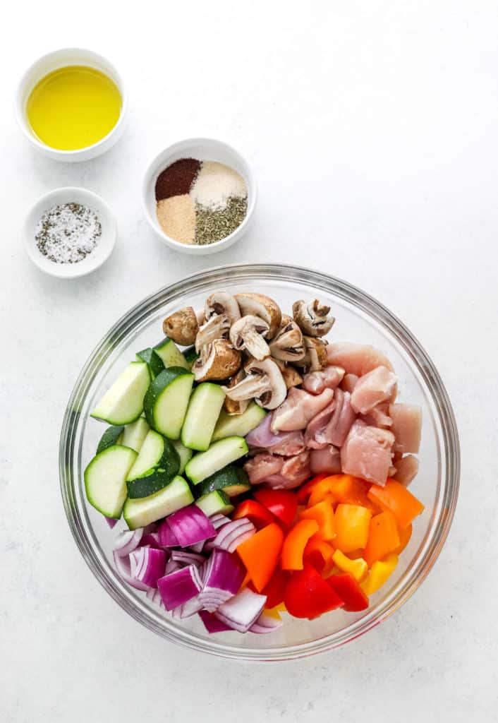 Round glass bowl filled with chopped peppers, purple onion, zucchini, mushrooms and chicken with a bowl of spices, bowl of oil and a bowl of salt and pepper behind it on a white surface.