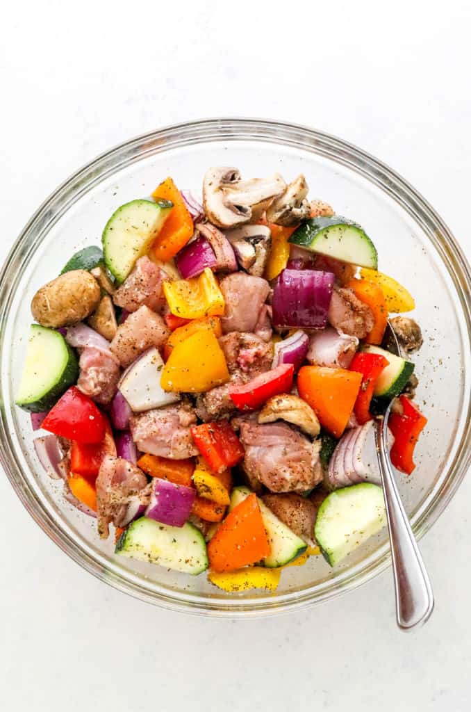 Round glass mixing bowl filled with uncooked, seasoned cut up chicken and veggies.