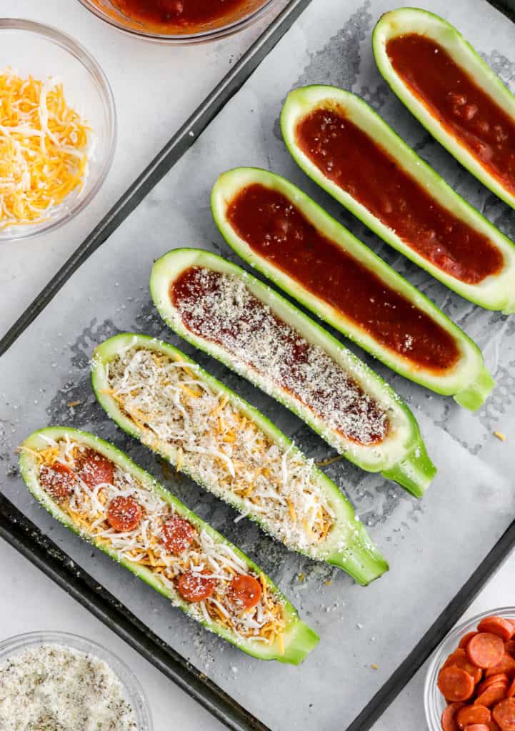 Zucchini pizza boats with cheese and pepperoni on a couple of them on top of a baking sheet with a bowl if shredded cheese next to it