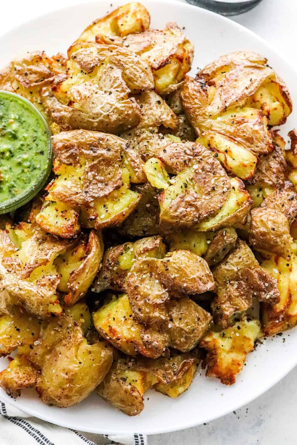 A pile of crispy cooked smashed baby potatoes on a round white plate with a small glass bowl of green pest sauce next to them on the plate.
