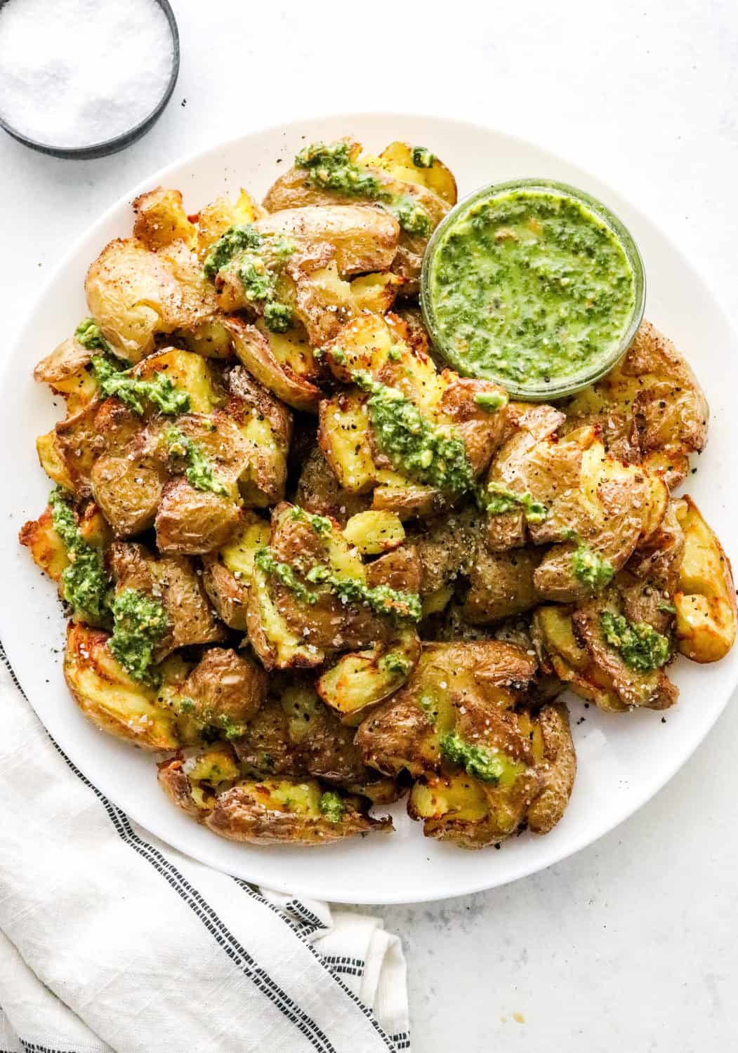 Pile of crispy round potatoes smashed on a round white plate drizzled with green sauce with a white and black striped linen next to it on a white surface. 