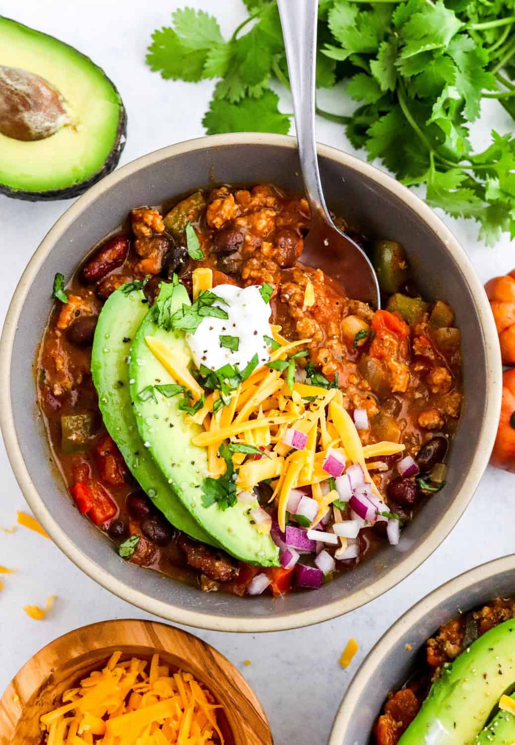 Round grey bowl of pumpkin chili topped with sliced avocado, shredded cheddar cheese, diced red onion an sour cream with another bowl of chili and a bowl of shredded cheddar cheese in front of it. 