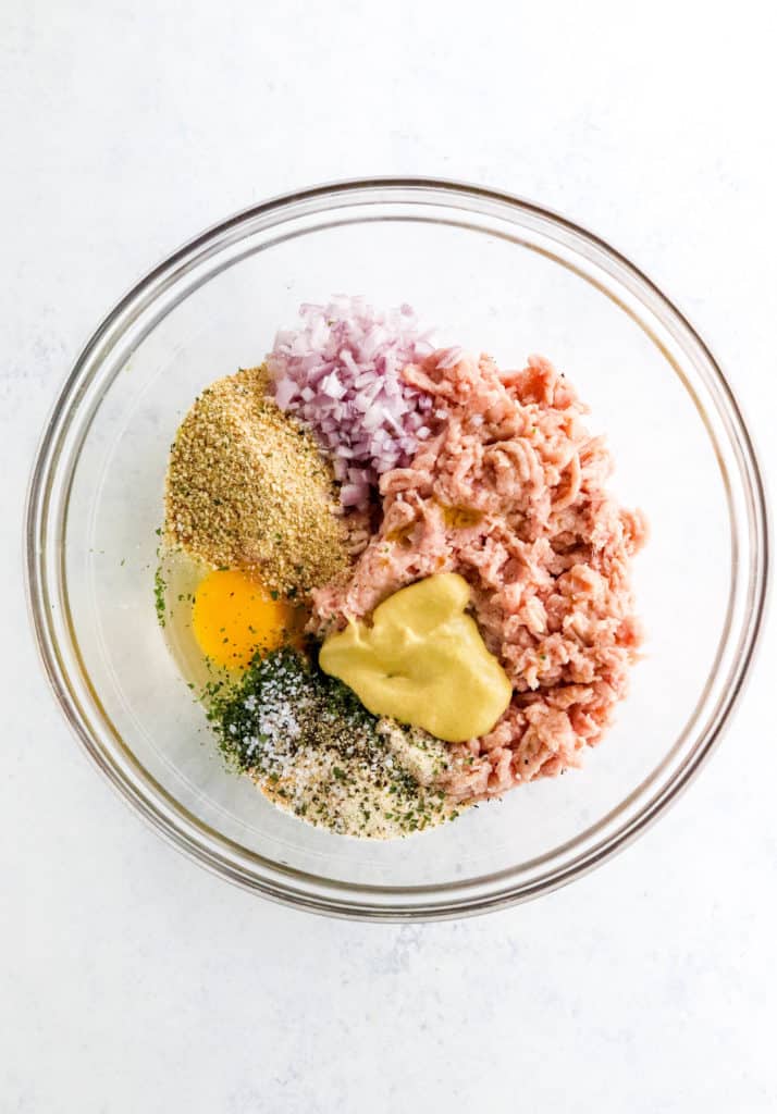 Round glass bowl with raw ground turkey, a cracked egg, breadcrumbs, mustard and diced shallots in it. 
