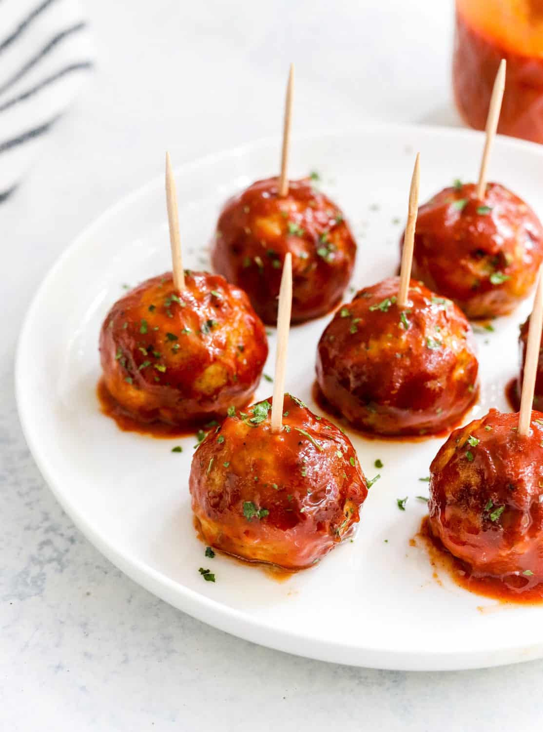 Crockpot BBQ Meatballs with toothpicks in the top of each meatballs on a round white plate with a striped towel and more bbq sauce behind it. 