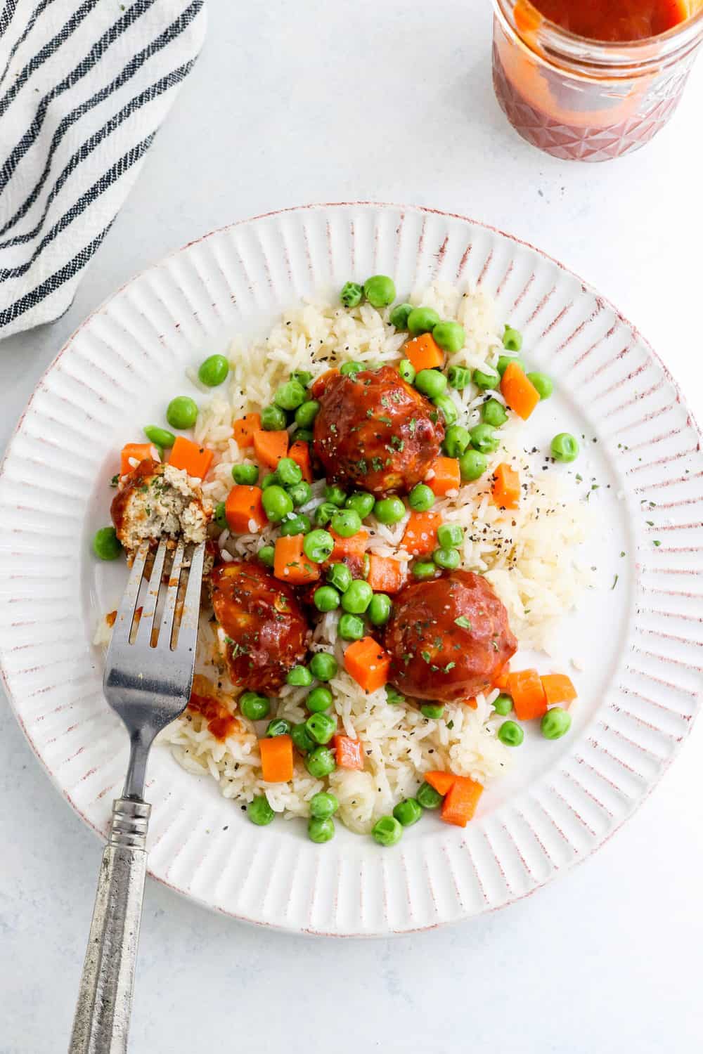 Round plate with white rice, mixed veggies and bbq meatballs on top of it with a fork cutting through one of the meatballs with a striped black and white towel and jar of bbq sauce behind it. 