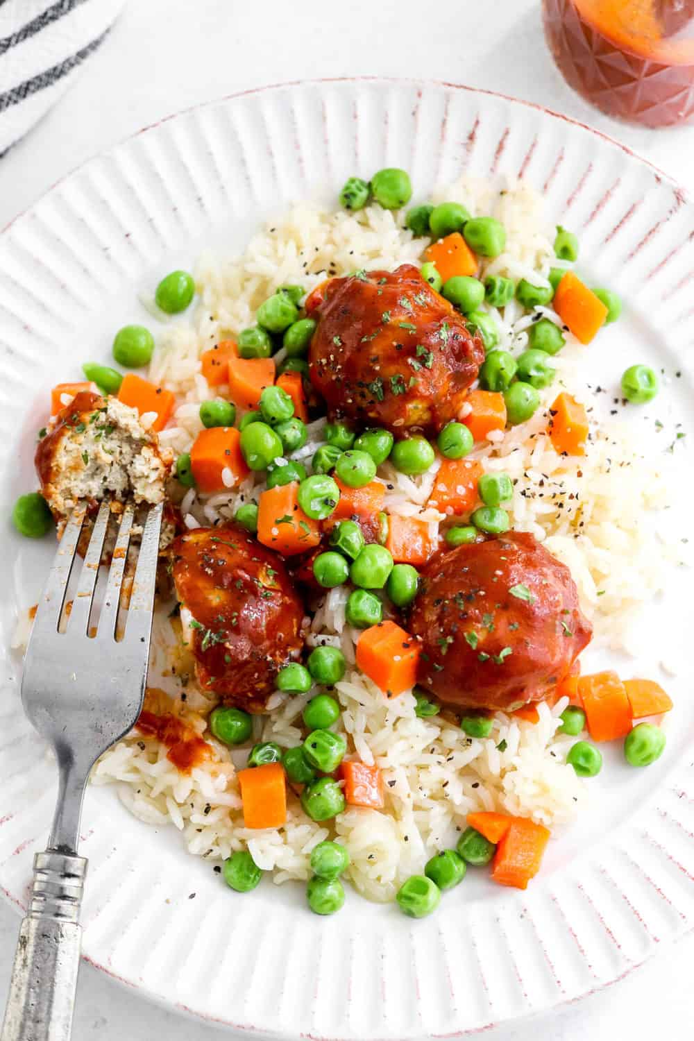 Textured plate filled with white rice, peas and carrots topped with sauce meatballs with a silver spoon on the plate. 