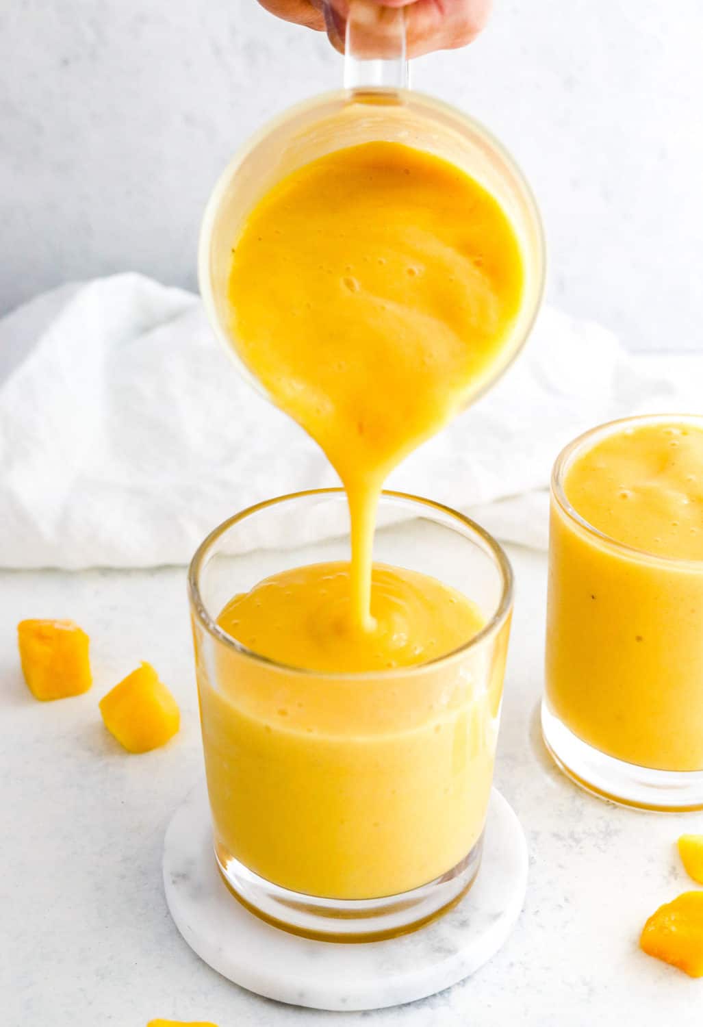 Hand pouring a pitcher of blended mango banana smoothie into a round glass with another glass filled with the smoothie mixture next to it and mango pieces around it. 