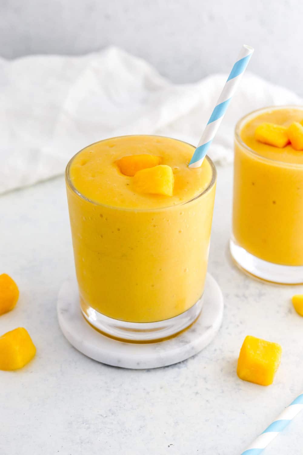 Glass fill with mango banana smoothie mixture with a blue and white stripped straw in the glass with pieces of frozen mango on top and more pieces of mango around the glass. 