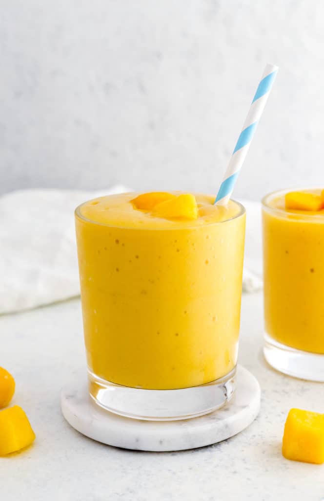 Round glass filled with yellow mango smoothie topped with a few pieces of mango with a blue and white straw in the glass and another glass filled with the Smoothie mixture next to it