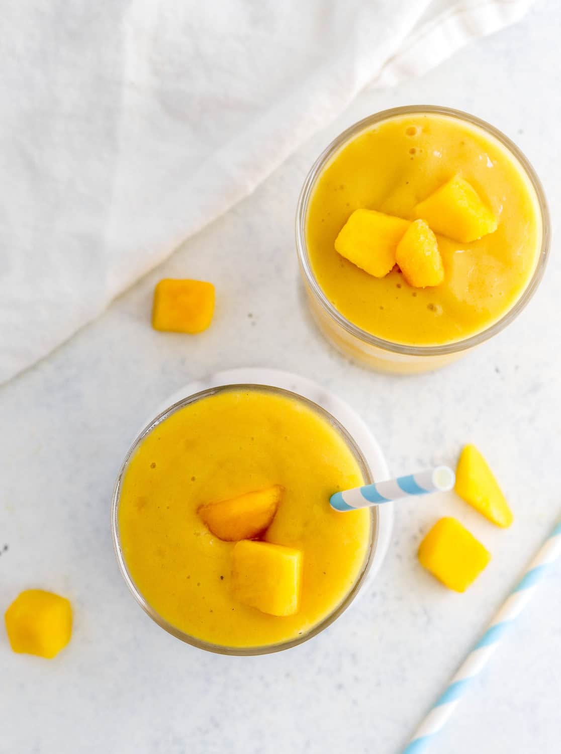 Overhead view of two mango banana smoothies in glasses with a blue and white straw in one of the glasses and pieces of cut mango around them. 
