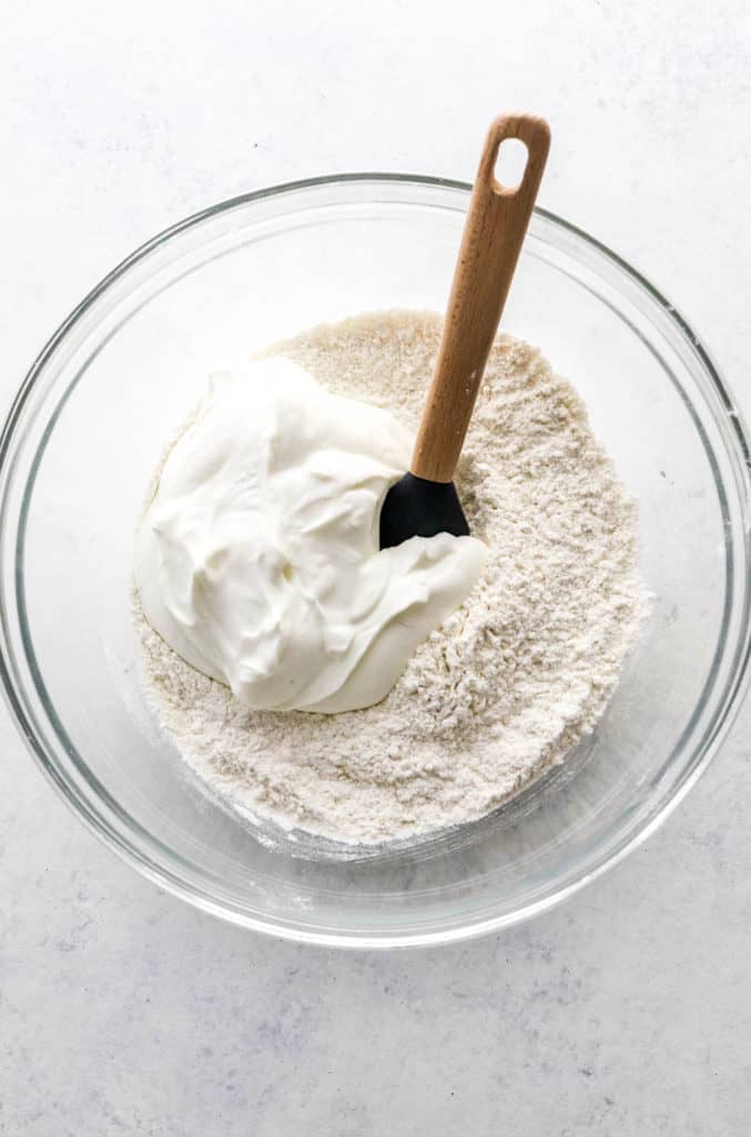 Flour and Greek yogurt in round glass mixing bowl with rubber spatula in it. 