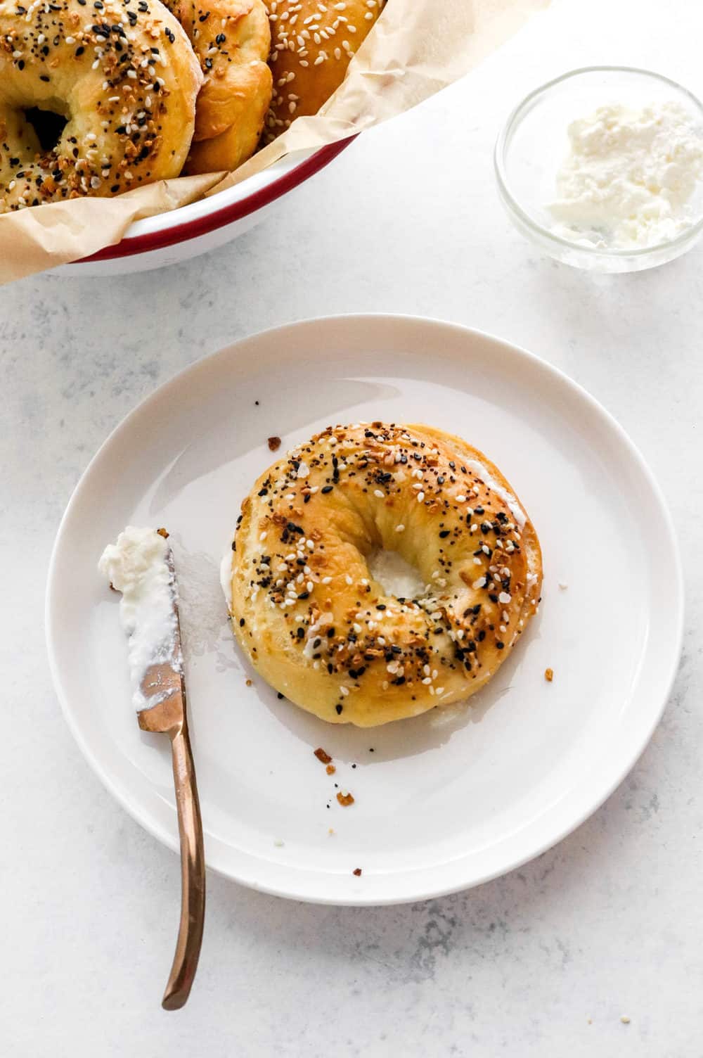 Single everything bagel on a plate with a knife with cream cheese on it next to it with a bowl of bagels behind it. 