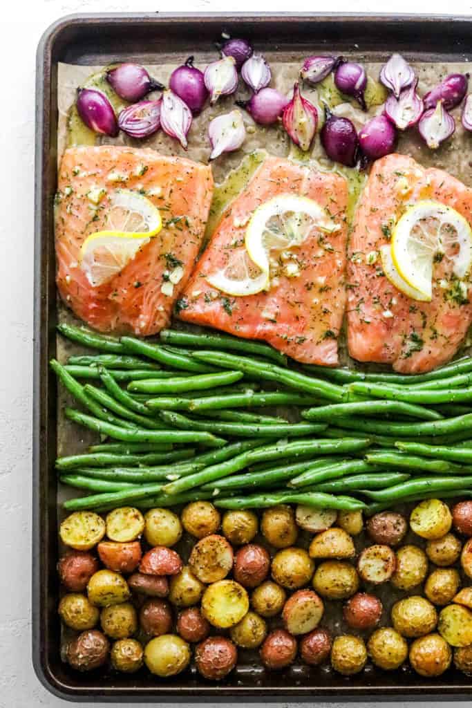 Sheet pan filled with onion, salmon filets with lemon slices on top of them, green beans and cut baby potatoes all separated on the pan