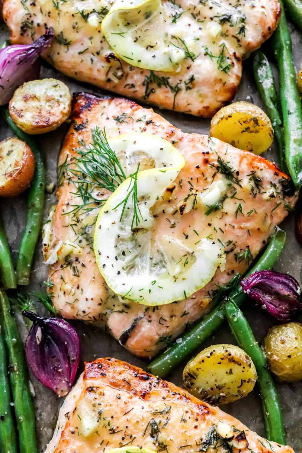 Close up of a cooked sheet pan salmon filet with a sliced lemon wedge on top of it with green beans, onion and potatoes around it. 