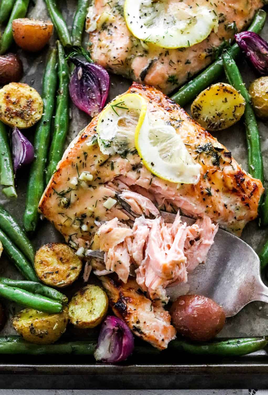 Close up of a cooked salmon filet being cut with a fork surrounded by green beans, purple pearl onions and crispy cooked baby potatoes. 