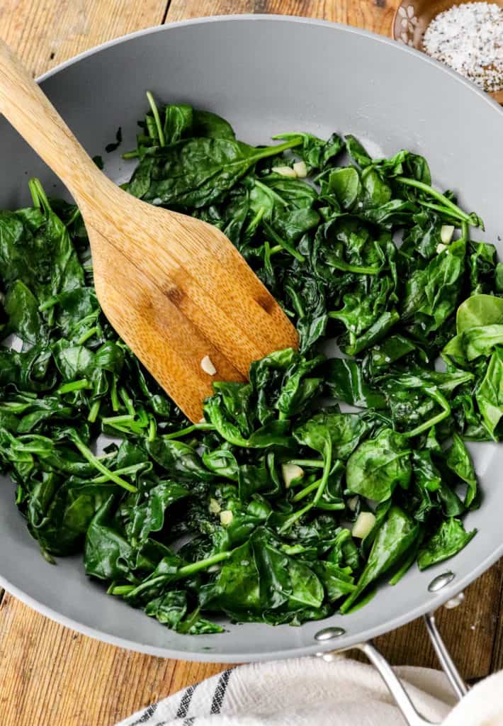 Spinach leaves cooked in a gray sauté pan with a wooden spoon in it