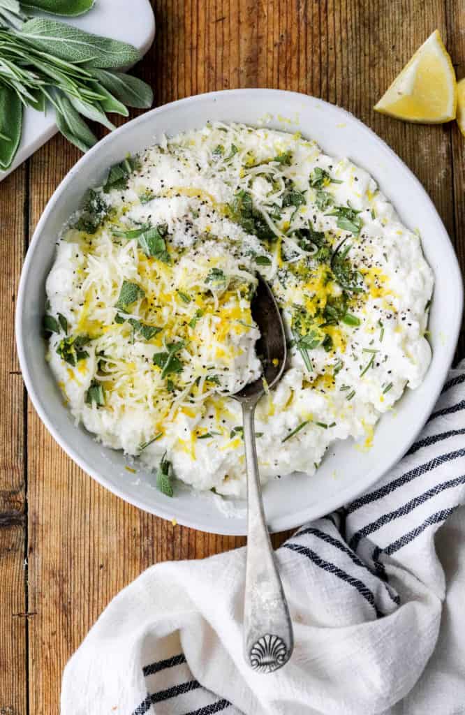 Round white bowl of ricotta cheese topped with herbs and lemon zest with a spoon in the bowl and more herbs on a cutting board behind it. 